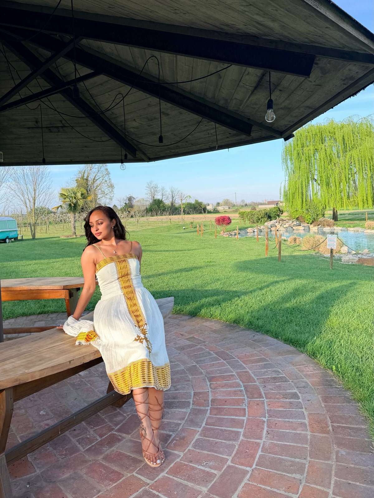 A woman wearing a white strapless Ethiopian National Dress with gold trim and lace-up strappy heels poses on a brick patio under a wooden pavilion, with a green lawn and trees in the background.