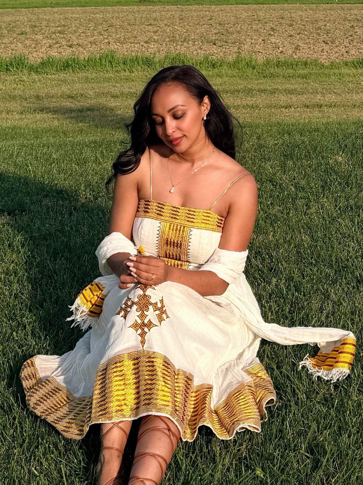 A woman sitting on a green grass field wearing a white Ethiopian National Dress with yellow and gold geometric patterns, a matching white shawl, and lace-up ankle boots.
