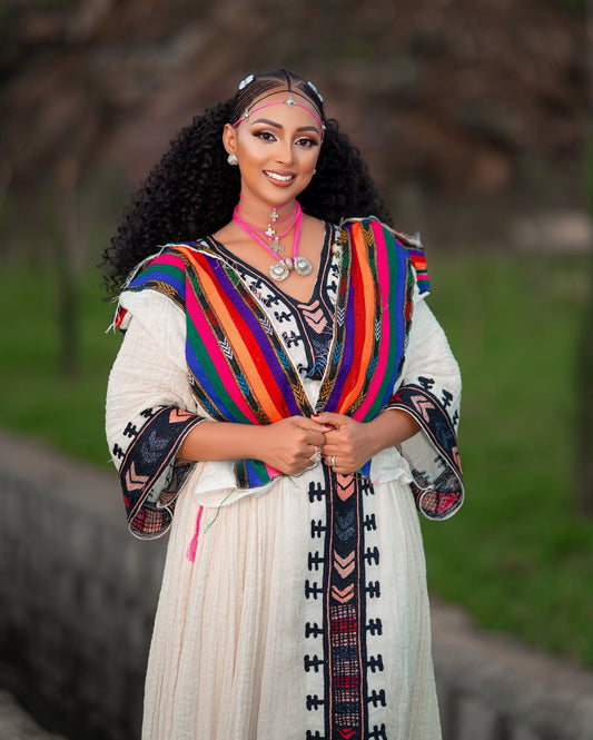 A woman wearing a traditional Ethiopian Axum Fetil Dress with a colorful striped shawl draped over her shoulders, standing outdoors against a blurred green and brown background.