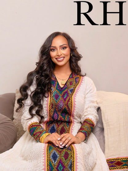 A woman with long, wavy dark hair sits on a beige couch wearing a white embroidered traditional  Axum Fetil  with colorful geometric patterns. The background is a plain beige wall with a black 'RH' logo in the top right corner.