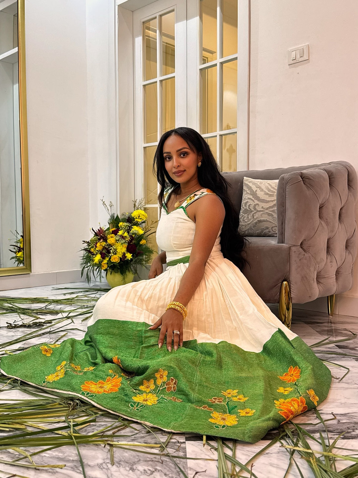 A woman sits on a marble floor in a living room setting, wearing a cream-colored traditional Ethiopian National Dress with a green skirt featuring yellow floral embroidery. A matching green embroidered dupatta is draped in front of her, with a gray tufted sofa and a vase of yellow flowers in the background.