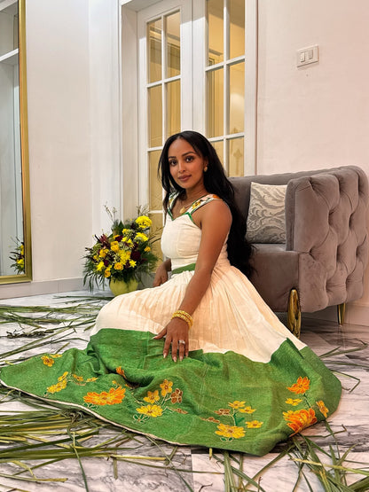 A woman sits on a marble floor in a living room setting, wearing a cream-colored traditional Ethiopian National Dress with a green skirt featuring yellow floral embroidery. A matching green embroidered dupatta is draped in front of her, with a gray tufted sofa and a vase of yellow flowers in the background.