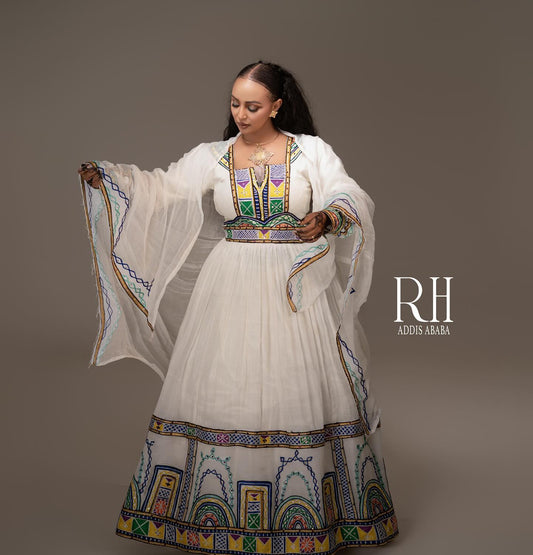 A woman modeling a traditional white Axum fetil  with colorful geometric patterns and sheer white sleeves, posed against a solid taupe background. The brand logo 'RH ADOIS ARABA' is visible on the right side.