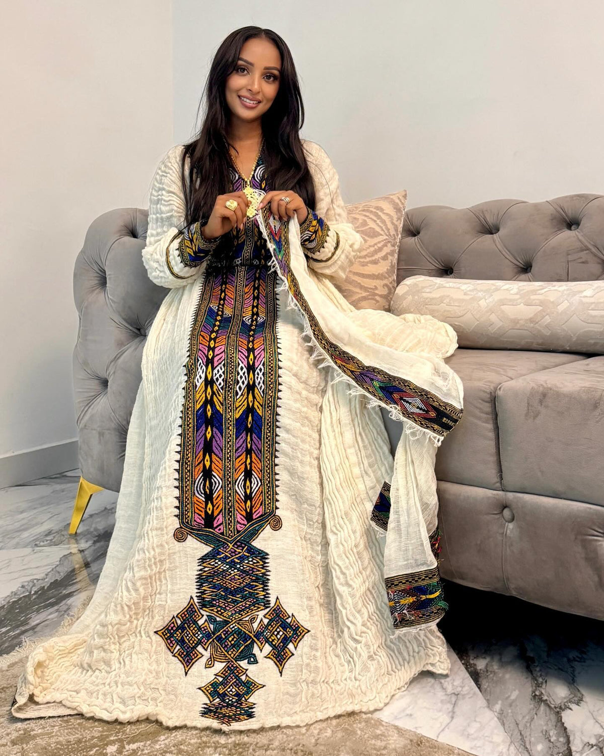 A woman wearing a long cream-colored embroidered traditional Ethiopian National Dress with a matching draped shawl, standing in front of a gray tufted sofa in a living room setting.