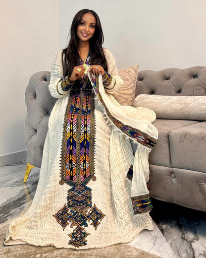 A woman wearing a long cream-colored embroidered traditional Ethiopian National Dress with a matching draped shawl, standing in front of a gray tufted sofa in a living room setting.