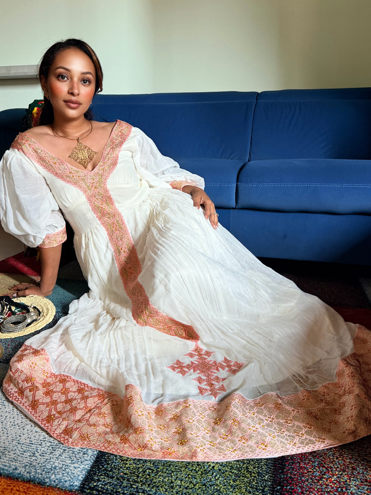 A woman wearing a long white maxi Ethiopian National Dress with peach-pink embroidered borders and a matching embroidered dupatta, sitting on a colorful patchwork rug in front of a blue sofa.