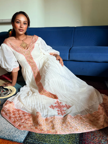 A woman wearing a long white maxi Ethiopian National Dress with peach-pink embroidered borders and a matching embroidered dupatta, sitting on a colorful patchwork rug in front of a blue sofa.