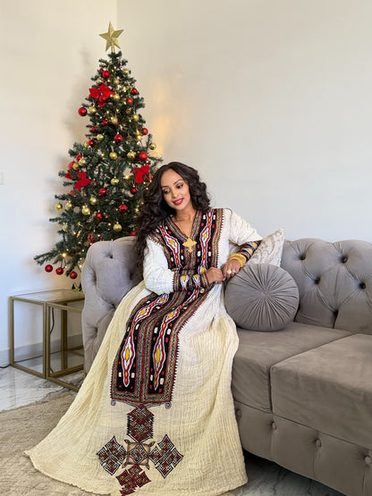 A woman wearing a long white Axum Fetil with intricate brown, red, and yellow embroidery sits on a gray tufted sofa next to a decorated Christmas tree.