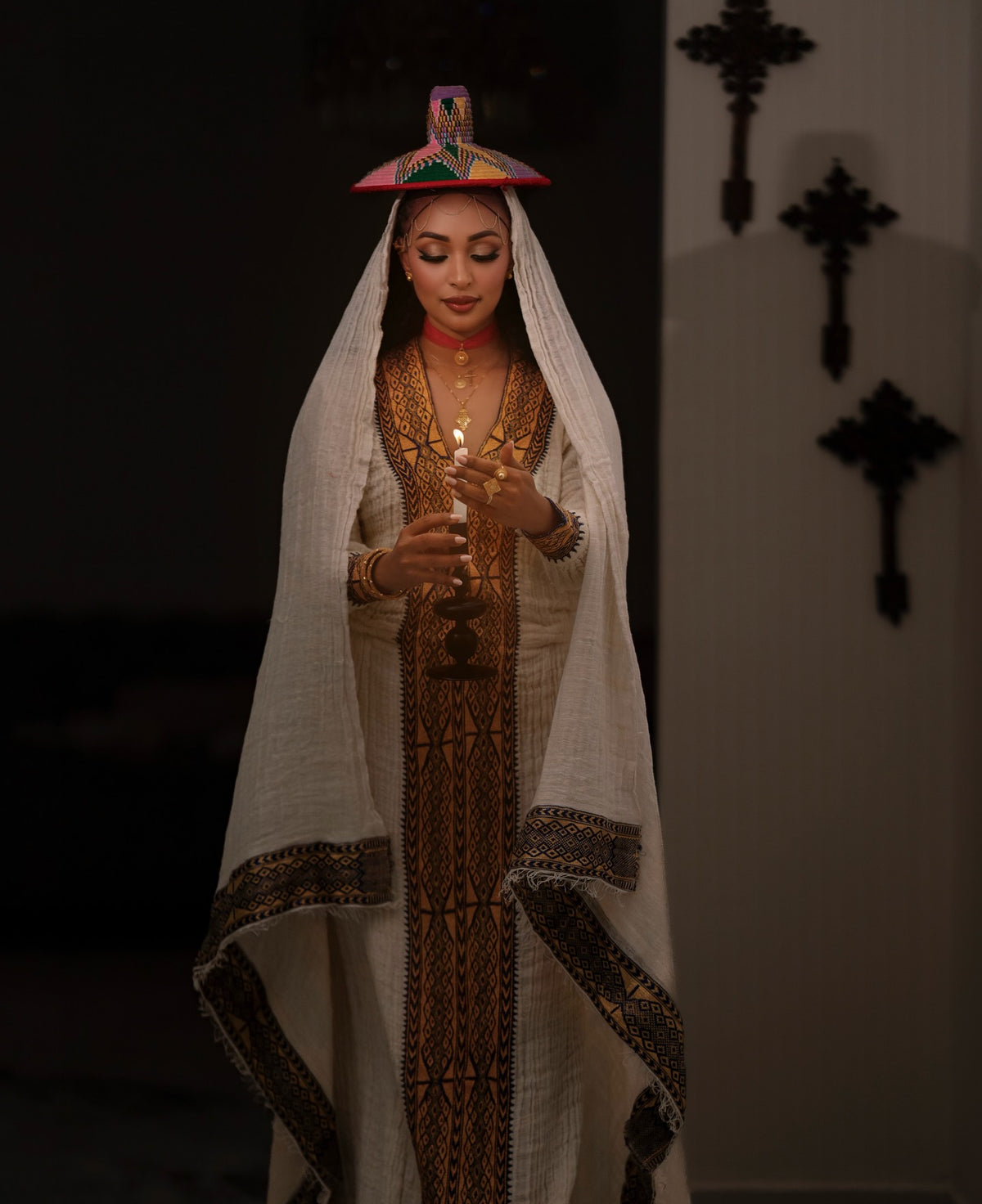 A woman wearing a traditional white embroidered Ethiopian National Dress with a matching veil and a small colorful hat, holding a candle in a dimly lit interior.