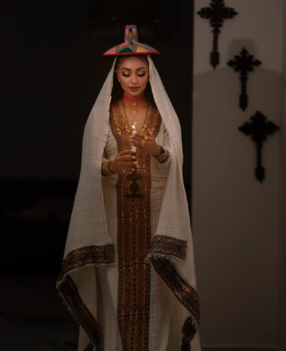 A woman wearing a traditional white embroidered Ethiopian National Dress with a matching veil and a small colorful hat, holding a candle in a dimly lit interior.