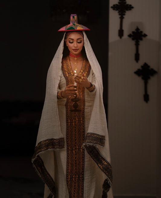 A woman wearing a traditional white embroidered Ethiopian National Dress with a matching veil and a small colorful hat, holding a candle in a dimly lit interior.