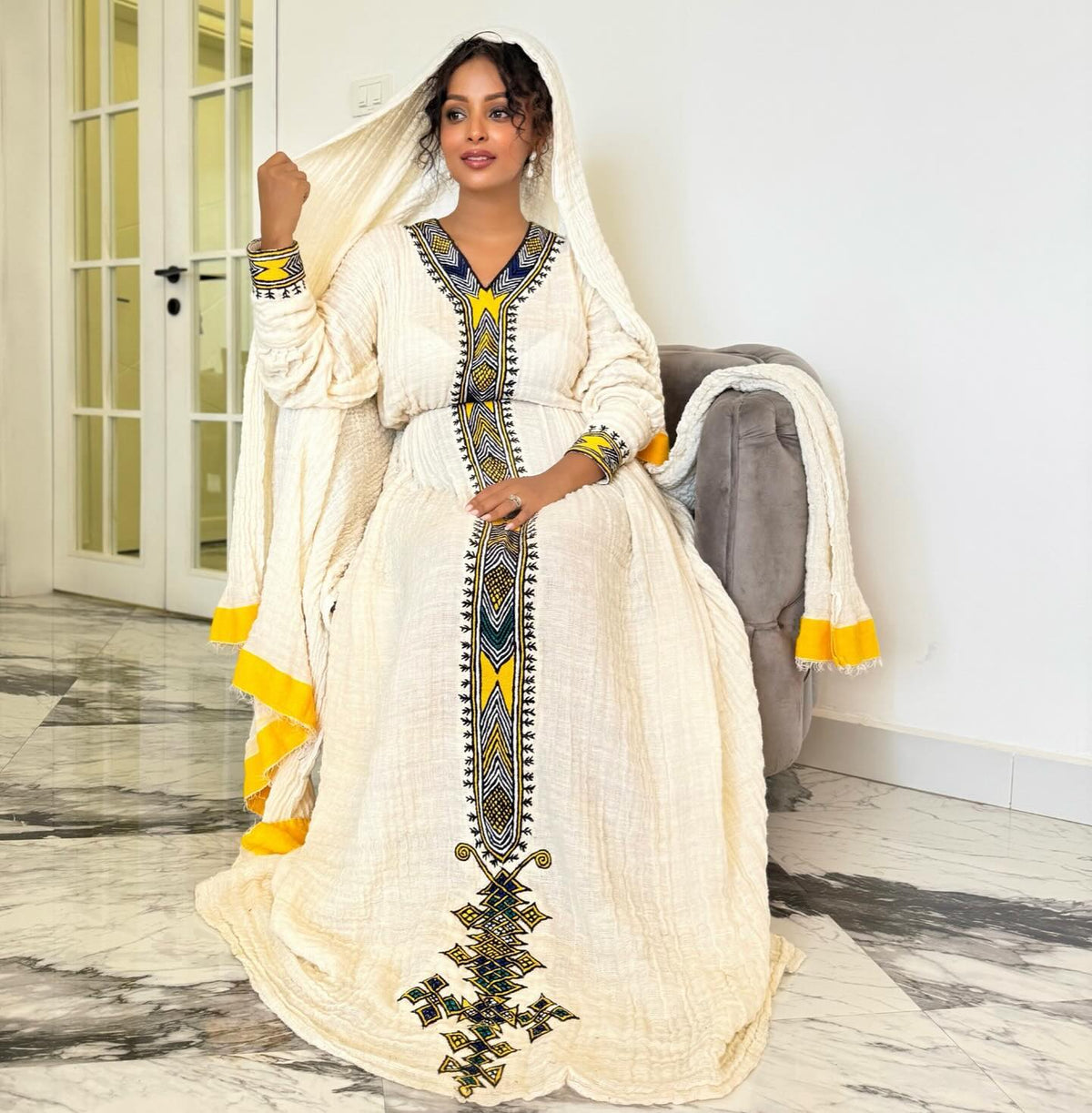 A woman models a traditional long white Ethiopian National Dress with yellow and blue geometric embroidery, holding a matching white veil with yellow fringe in an indoor setting.