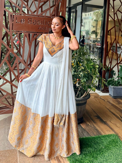 A woman models a white Ethiopian National Dress  with gold embroidery on the bodice and a gold patterned border on the skirt, set against a decorative wooden screen and potted plants.