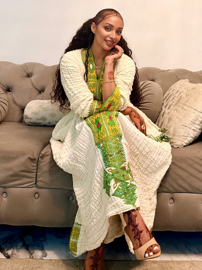 A woman sits on a tufted gray sofa wearing a long cream knit cardigan draped over a green and yellow printed Axum Fetil, with patterned sandals, in an indoor setting.