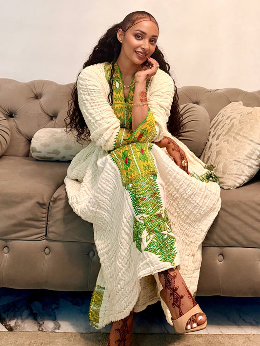 A woman sits on a tufted gray sofa wearing a long cream knit cardigan draped over a green and yellow printed Axum Fetil, with patterned sandals, in an indoor setting.