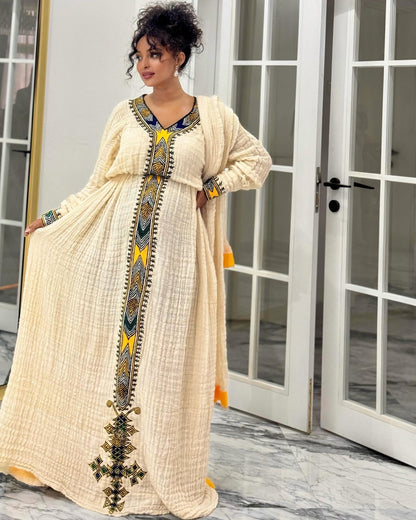 A woman models a long cream-colored textured Ethiopian National Dress  with colorful geometric embroidery in yellow, blue, and black along the neckline, center front, and hem, standing in front of white glass doors.