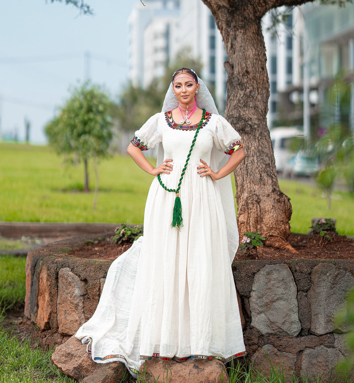 A woman wearing a traditional white Axum Fetil with colorful embroidered details, a green tassel necklace, and a veil, posing outdoors next to a tree.
