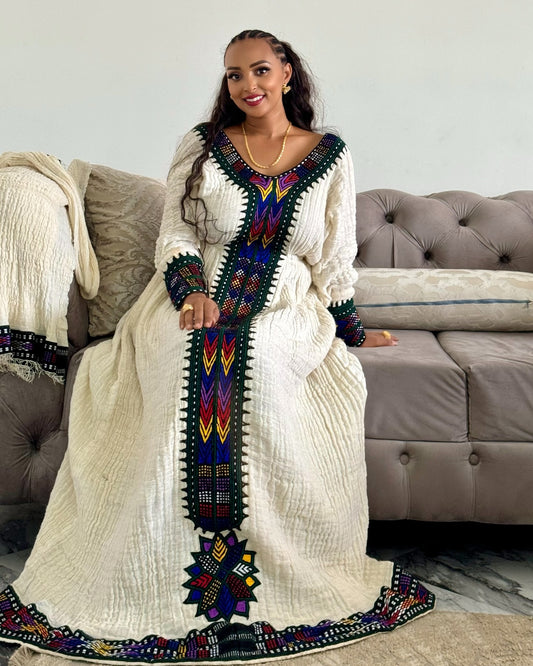 A woman wearing a long cream-colored traditional Ethiopian National Dress with colorful geometric embroidery, sitting on a tufted sofa in a living room setting.