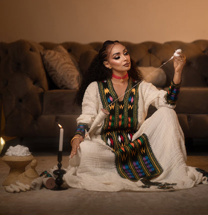 A woman sits cross-legged on a carpeted floor in front of a tufted brown sofa, wearing a cream-colored traditional Axum Fetil with colorful geometric embroidery. She holds a white flower near her face, with a lit candle in a black holder and a small bowl of white flowers beside her.