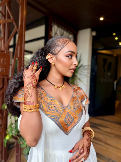 A woman wearing a white Ethiopian National Dress  with an orange and gray geometric embroidered neckline, styled with gold jewelry and henna on her hands, standing in a modern indoor setting with wooden columns.