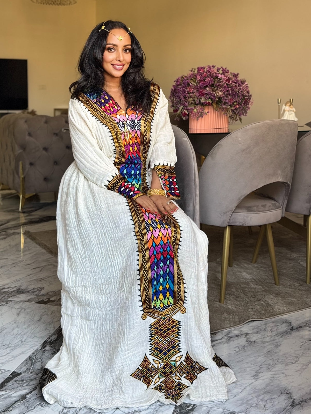 A woman wearing a white long-sleeve Axum Fetil  dress with vibrant multicolor geometric embroidery and gold trim, seated in a modern living room setting.