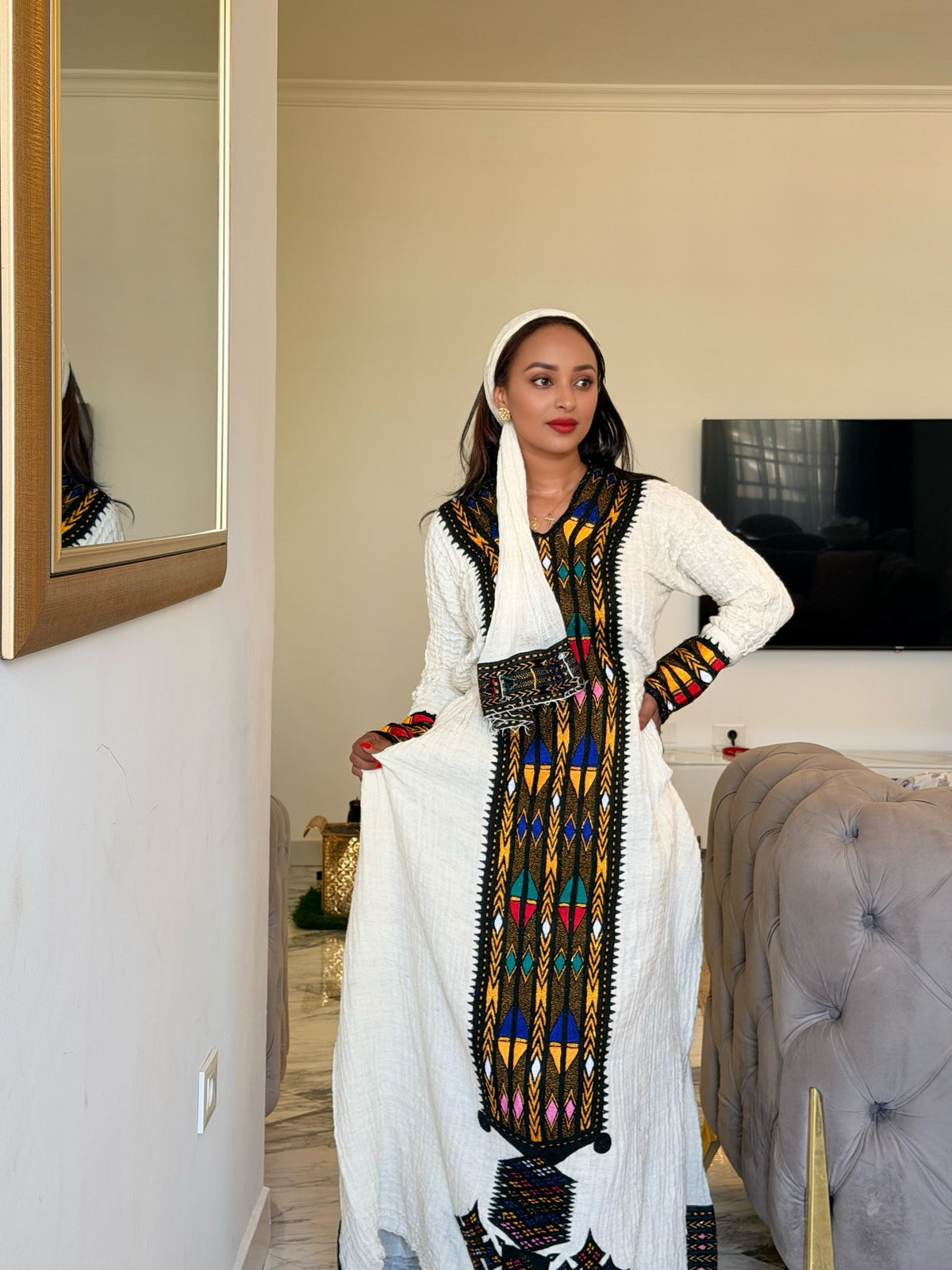 A woman modeling a traditional white Ethiopian National Dress with colorful geometric embroidery and a black waist belt, standing in a bedroom setting.