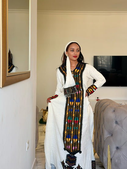 A woman modeling a traditional white Ethiopian National Dress with colorful geometric embroidery and a black waist belt, standing in a bedroom setting.