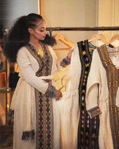 A woman with dark curly hair stands beside a clothing rack, holding a cream-colored Axum Fetil with a black, blue, and multicolor geometric embroidered border. Other similar dresses are hanging on the rack behind her in a softly lit indoor setting.