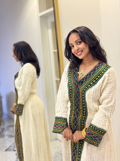 A woman wearing a cream-colored textured Axum Fetil with green, yellow, and red geometric embroidery, standing in front of a mirror that shows her back view.