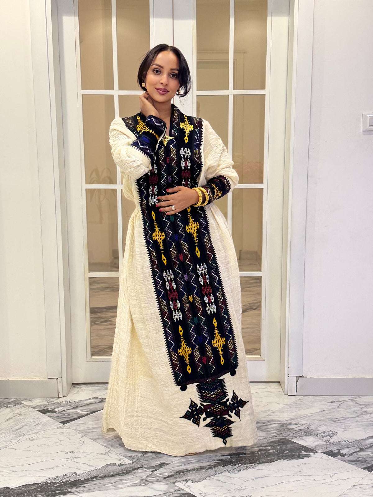 A woman wearing a long cream-colored traditional Axum Fetil with a large black embroidered panel featuring colorful geometric patterns, standing in front of a glass-paneled door on a marble floor.