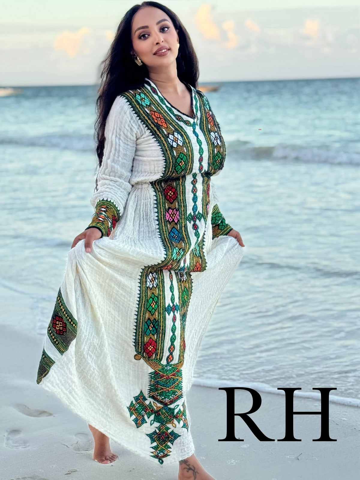 A woman stands on a beach wearing a long white Axum Fetil with colorful embroidered patterns in green, red, and blue. The background shows the ocean and sky, and a large 'RH' watermark is visible in the bottom right corner.