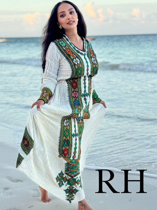 A woman stands on a beach wearing a long white Axum Fetil with colorful embroidered patterns in green, red, and blue. The background shows the ocean and sky, and a large 'RH' watermark is visible in the bottom right corner.