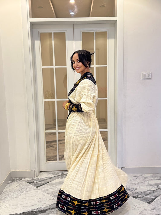 A woman wearing a long cream-colored traditional Axum Fetil with black and gold patterned trim, standing in front of a glass-paneled door in an indoor setting.
