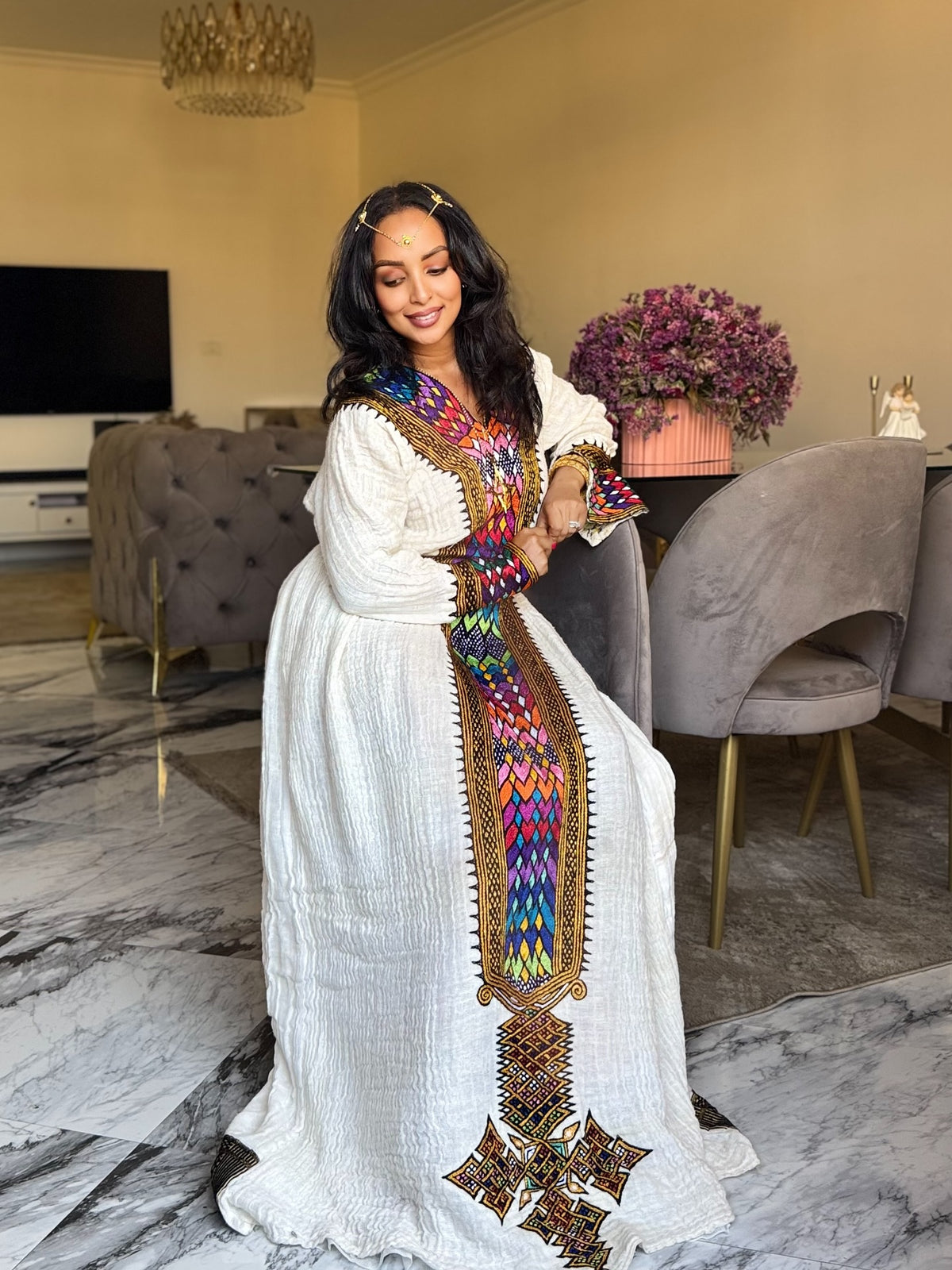 A woman wearing a long white Axum Fetil  with vibrant multicolor geometric embroidery and a gold-trimmed border, standing in a modern living room with gray velvet chairs and a marble floor.
