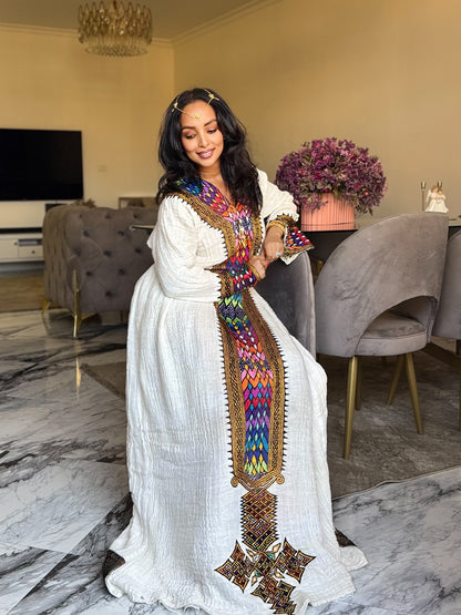 A woman wearing a long white Axum Fetil  with vibrant multicolor geometric embroidery and a gold-trimmed border, standing in a modern living room with gray velvet chairs and a marble floor.