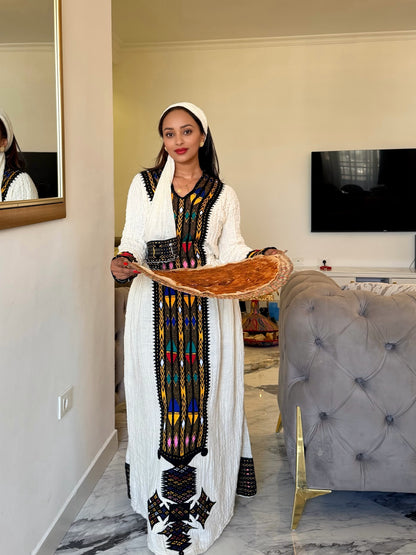 A woman wearing a long white traditional Ethiopian National Dress with colorful geometric embroidery and a matching headscarf, holding a large flatbread in a modern living room setting.