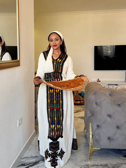 A woman wearing a long white traditional Ethiopian National Dress with colorful geometric embroidery and a matching headscarf, holding a large flatbread in a modern living room setting.