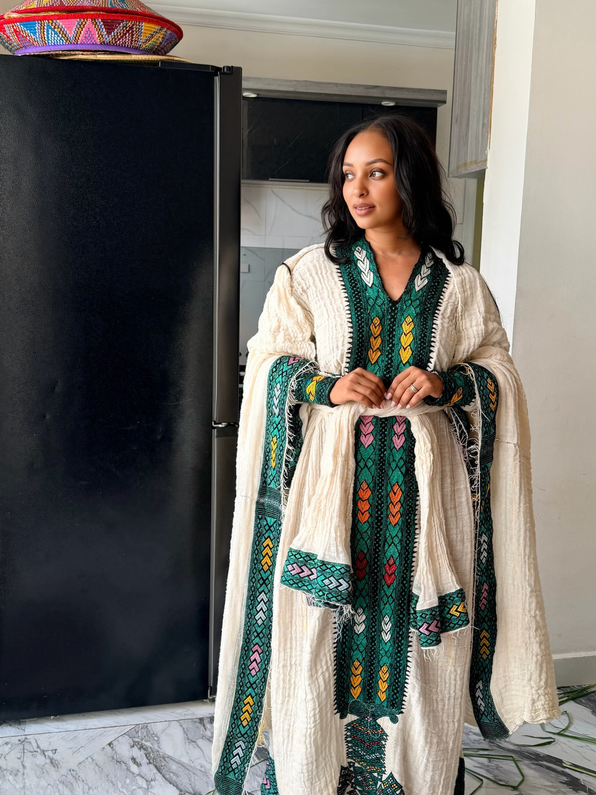 A woman wearing a cream-colored textured Axum Fetil with green, yellow, and red geometric embroidery, standing in a home interior next to a black refrigerator.