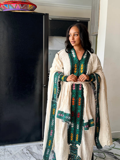 A woman wearing a cream-colored textured Axum Fetil with green, yellow, and red geometric embroidery, standing in a home interior next to a black refrigerator.