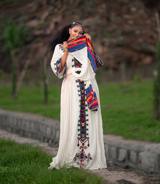 A woman outdoors wearing a long cream Axum Fetil Dress with colorful geometric embroidery and holding a matching striped textile in front of her face. The background shows a blurred natural setting with green grass and a stone wall.