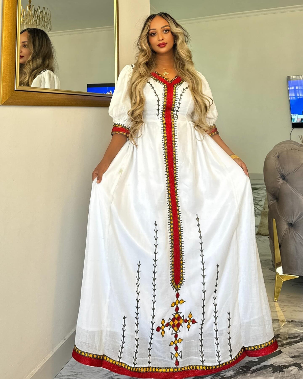 A woman models a traditional white Ethiopian National Dress with red and green embroidered details, standing in an indoor room with a mirror and a gray tufted chair visible in the background.