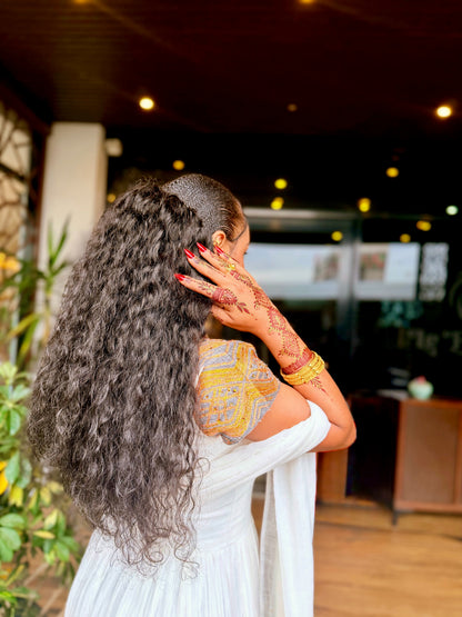 A woman with very long, dark, curly hair is shown from behind, wearing a Ethiopian National Dress  and displaying intricate red henna designs on her hand. She is standing in a dimly lit indoor space with warm bokeh lights in the background.