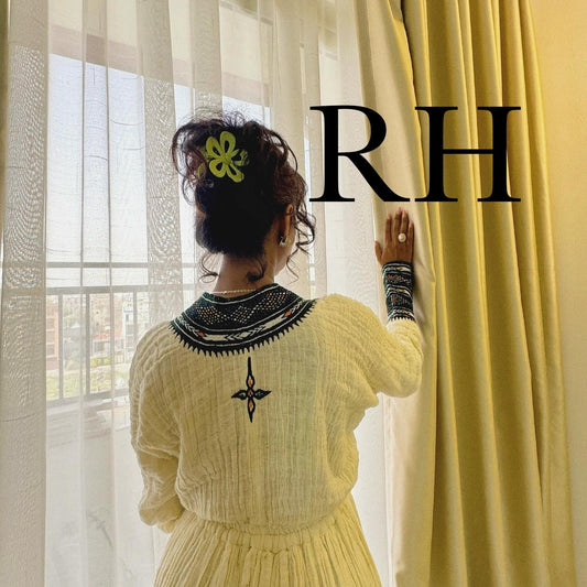A woman viewed from behind wearing a cream-colored textured Ethiopian National Dress with black embroidered detailing around the collar and on the back, standing by a window next to yellow curtains. A large black 'RH' text overlay is visible on the right side.