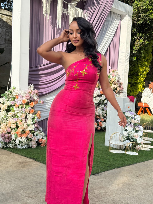 A woman wearing a bright fuchsia pink one-shoulder evening Ethiopian National Dress with gold embroidery on the bodice, standing in front of a decorated event backdrop with purple drapes and floral arrangements.