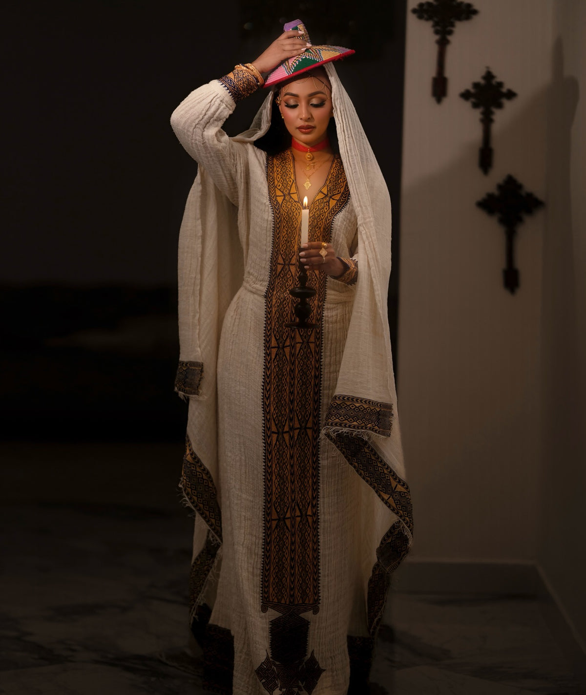 A woman wearing an elegant traditional Ethiopian National Dress with a long cream-colored embroidered robe, a matching head covering, and a patterned dress, holding a lit candle. She is posed in a dimly lit interior with a dark wall and hanging cross decorations in the background.