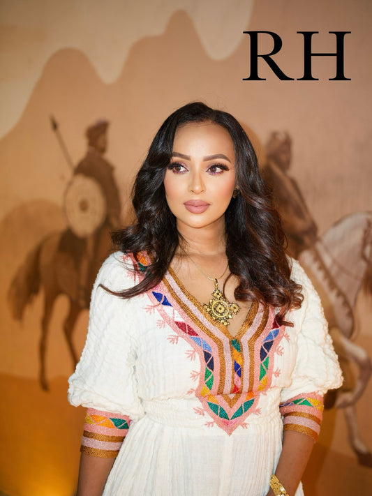 A woman with long dark wavy hair is wearing a white embroidered Ethiopian National Dress with colorful geometric patterns around the neckline and sleeve cuffs. She is standing in front of a warm beige backdrop with silhouettes of riders on horseback and the letters 'RH' in the top right corner.