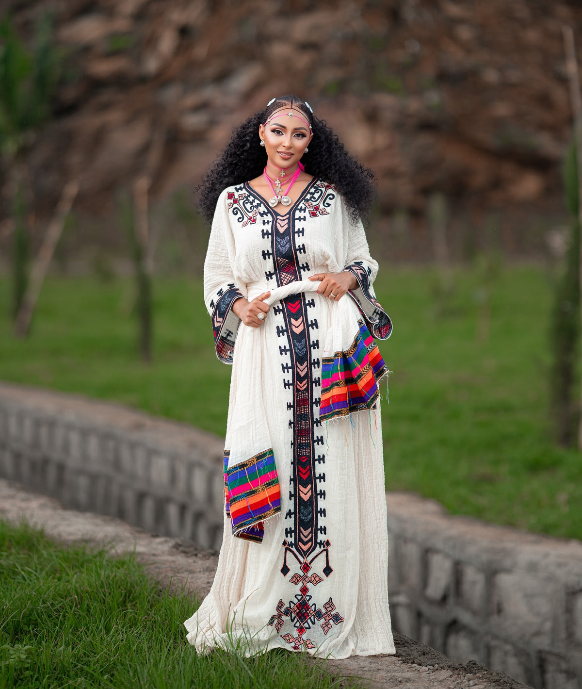 A woman stands outdoors wearing a long white embroidered Axum Fetil Dress with colorful geometric patterns and a matching headpiece. The background shows grass, trees, and a stone wall.