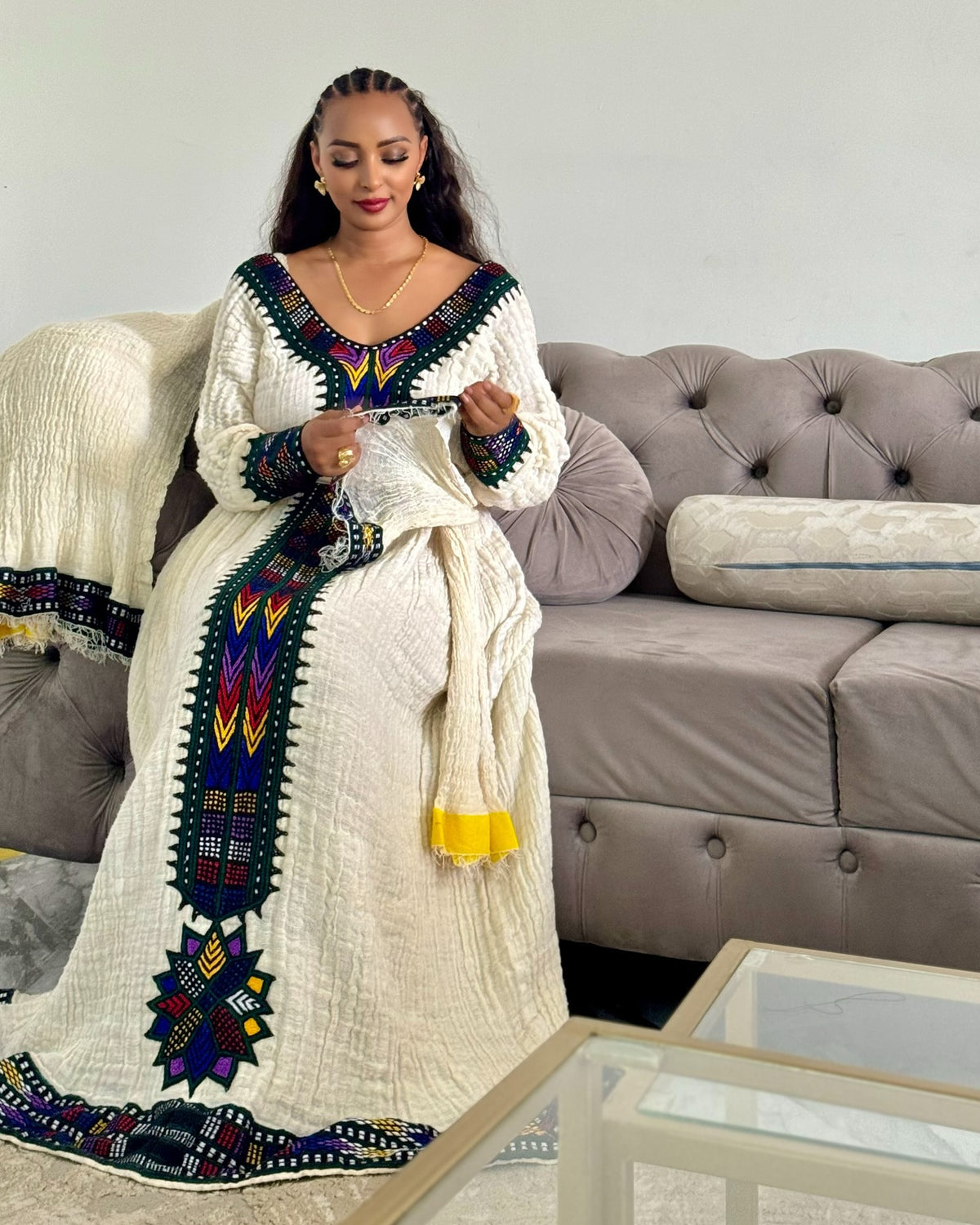 A woman wearing a long white textured Ethiopian National Dress with colorful geometric embroidery in green, blue, yellow, and red, sitting on a gray tufted sofa.