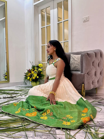 A woman wearing a traditional Indian salwar Ethiopian National Dress sits on a marble floor in a decorated room. The dress has a cream-colored upper bodice and a vibrant green skirt decorated with yellow floral embroidery.