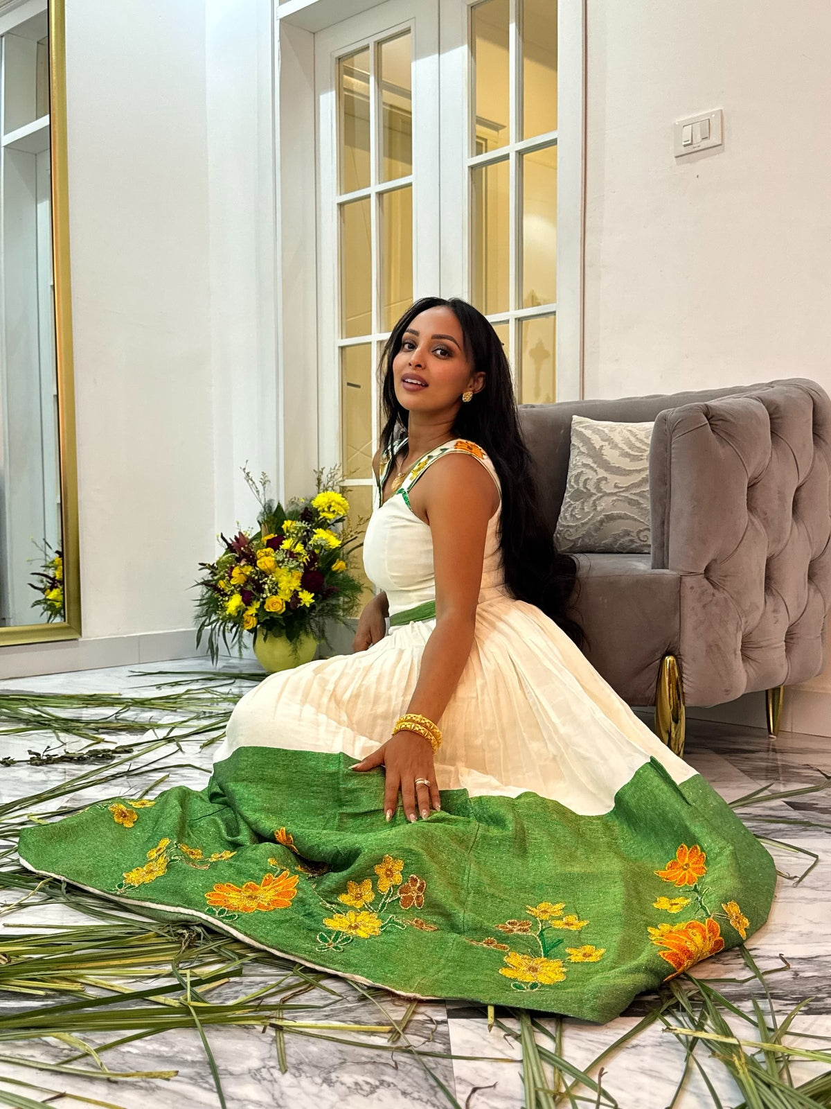 A woman wearing a traditional Ethiopian National Dress sits on a marble floor in a decorated room. The outfit consists of an off-white sleeveless kurta with green and gold embroidery, paired with a matching off-white skirt and a green dupatta featuring yellow floral patterns.